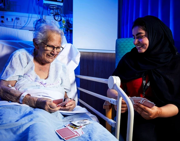 Royal Voluntary Service On Ward volunteer playing cards with a hospital patient who is in their bed